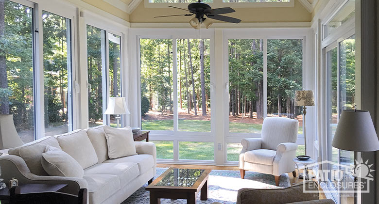 Interior of a white sunroom with white upholstered couch and chair, coffee table and lamps looking out to the woods beyond.