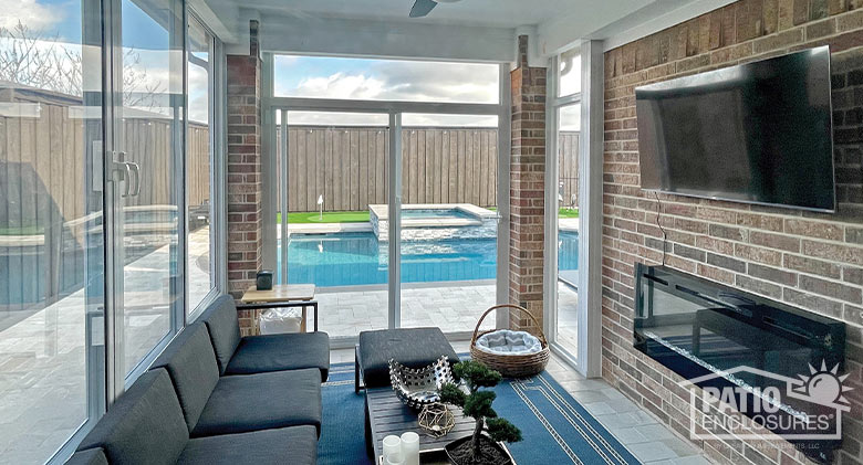 A glass patio enclosure with couch, wall-mounted TV and fireplace and view of pool and putting green.
