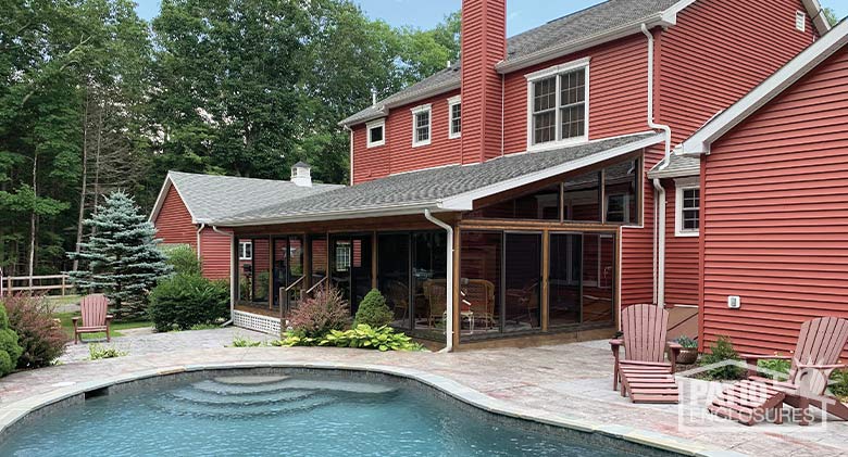 A pool and patio in the foreground with a glass-enclosed porch attached to a two-story red home with nicely landscaped yard.
