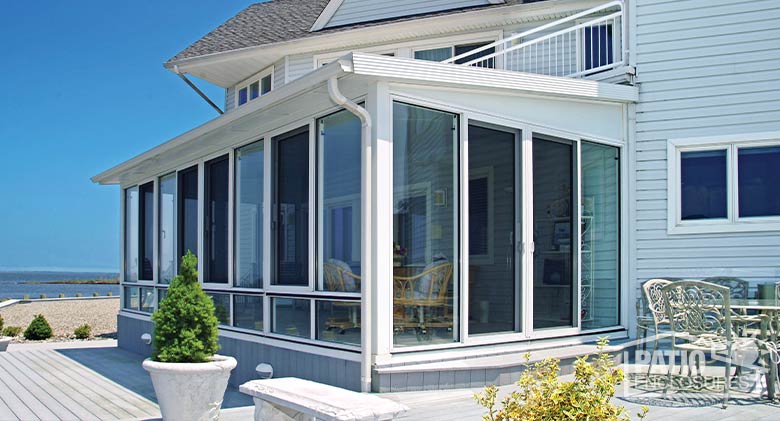 A simple, white sunroom with shed roof on a gray house surrounded by a wood deck and view of the beach and water beyond.