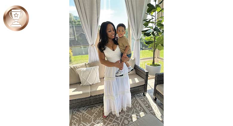 Woman in a white dress holding a small boy wearing a t-shirt and shorts standing in a sunroom with drapes tied back.