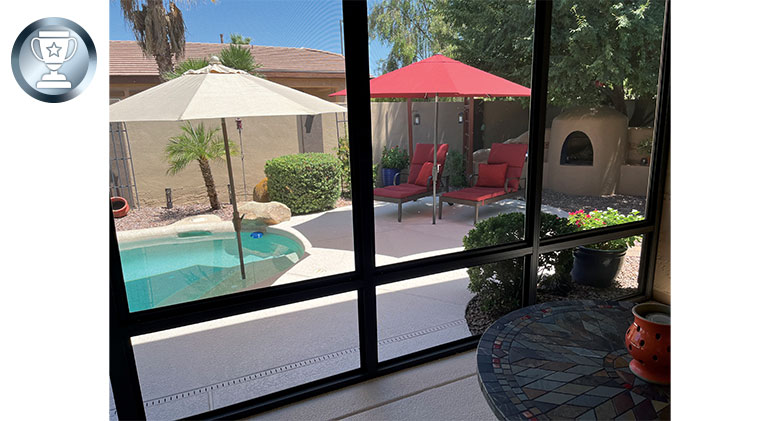 View of a pool, umbrellas, red chaise lounges and outdoor fireplace from the interior of a sunroom.