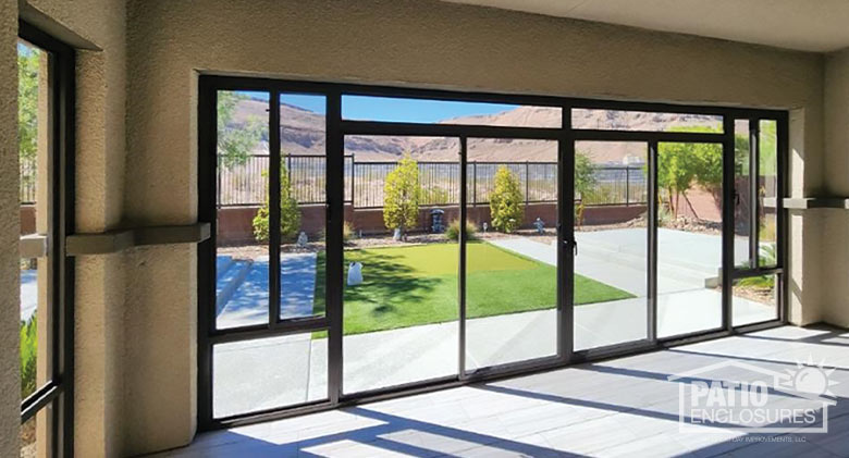The interior of a glass-enclosed patio gives us a view of the backyard with some small trees and mountains beyond.