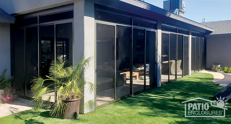 A brown-framed screen room encloses a corner patio in a gray stucco home. Astro-turf and plants surround the room.