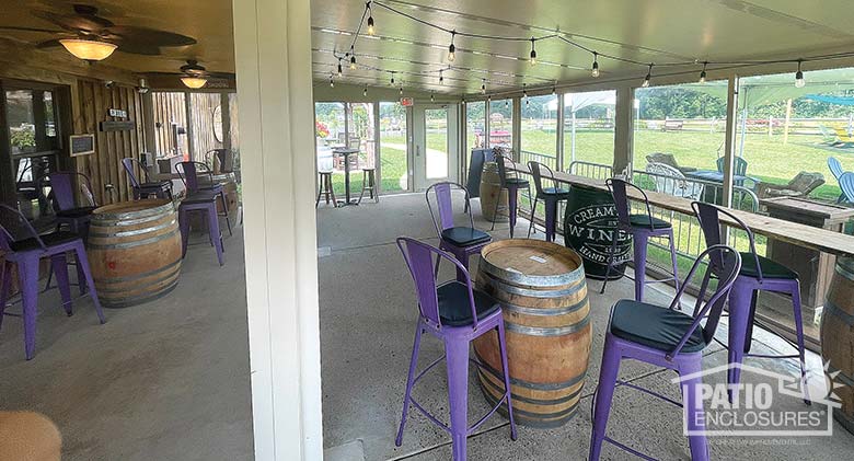 Sunroom interior of a winery with wine barrel tables and purple chairs, concrete floor and lights on the ceiling.