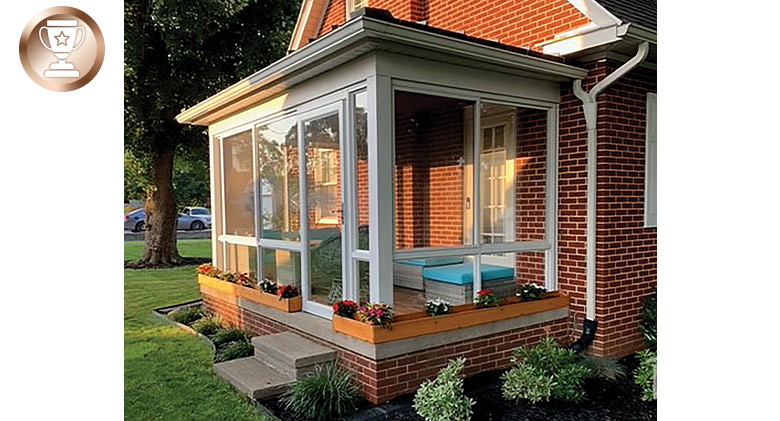A glass-enclosed porch on a brick house with flowers in window boxes around it and manicured flower beds with small shrubs.