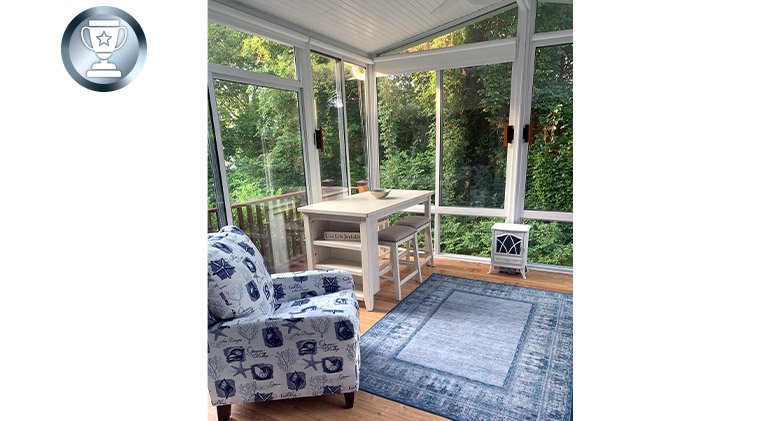Interior of a sunroom with gable roof, comfortable chair in blue and white print, blue rug and small writing desk.