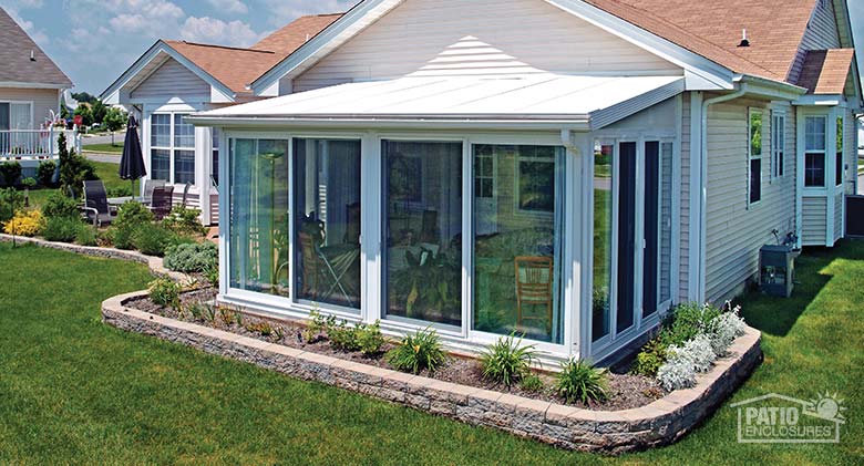 A white glass sunroom with a shed roof on the back of a house, flowerbed with low paver wall surrounds the room.