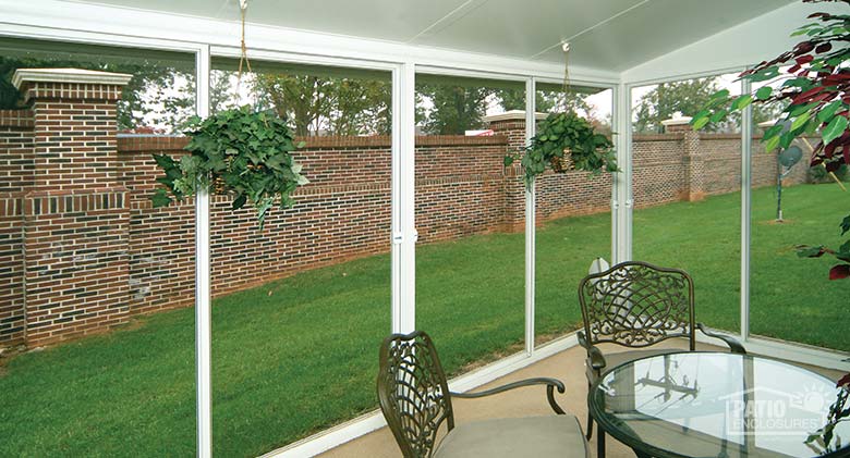 A Interior of a screen room with a table and chairs, hanging plants and a view of the lawn and a brick wall outside.