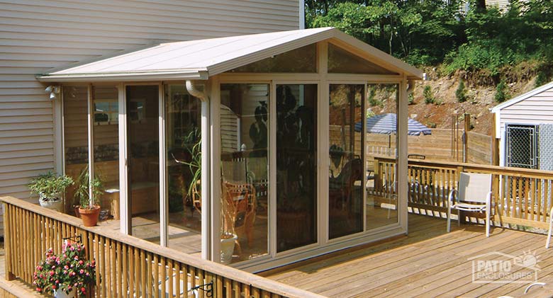Exterior of a tan sunroom with gable roof built on a wooden deck with railing and pink flowers on the left.