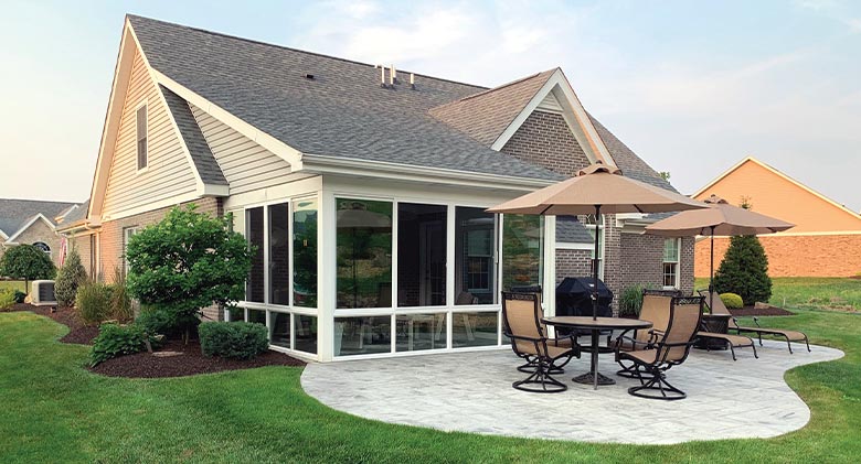 A beautiful patio enclosure on the back of a home with a table, chairs and umbrellas on a large patio in front of it.