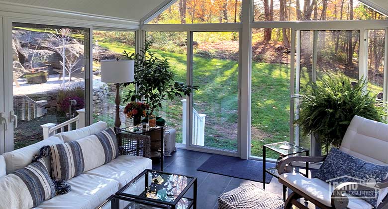 Gable roof sunroom interior with tile floor, comfortable couch and chair, and glass coffee table. View of woods/autumn trees.