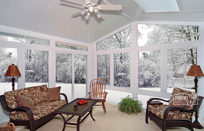 Sunroom interior with ceiling fan, indoor/outdoor furniture with patterned cushions and view of snow-covered landscape.