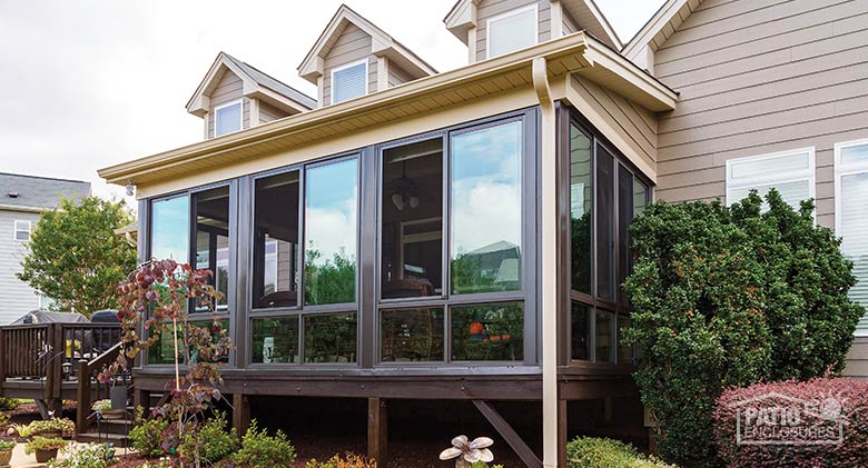Glass deck enclosure with brown frame on tan house with dormers. Nice landscaping and bushes surround the room.