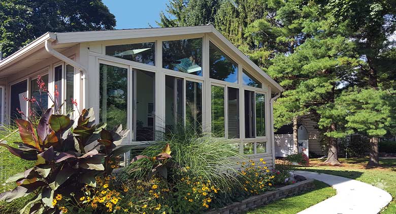 Large tan sunroom with gable roof and lush plantings of yellow flowers and grasses in front of it, pine trees as backdrop.