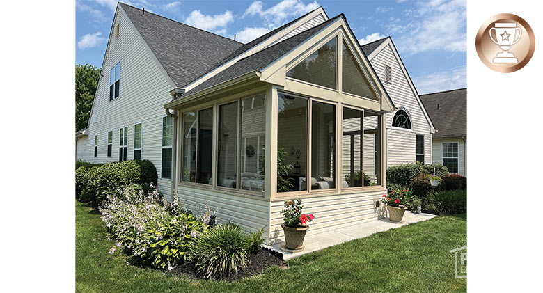 A beige house with a glass-enclosed patio with gable roof and a solid, sided knee wall. Nicely landscaped around sunroom.