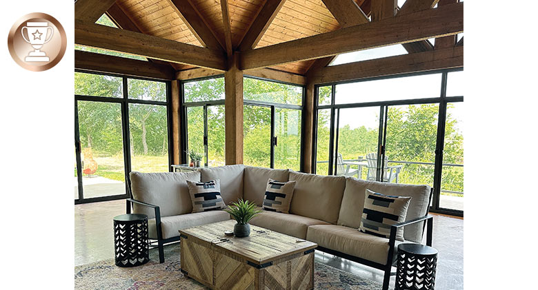 The interior of a glass sunroom featuring a soaring ceiling with wood beams and comfortable seating area.