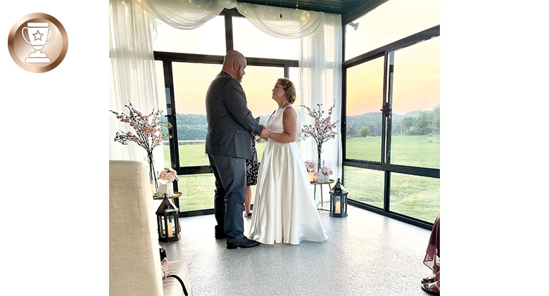 Bride and groom facing each other and holding hands. Through the sunroom glass you can see the sun setting.
