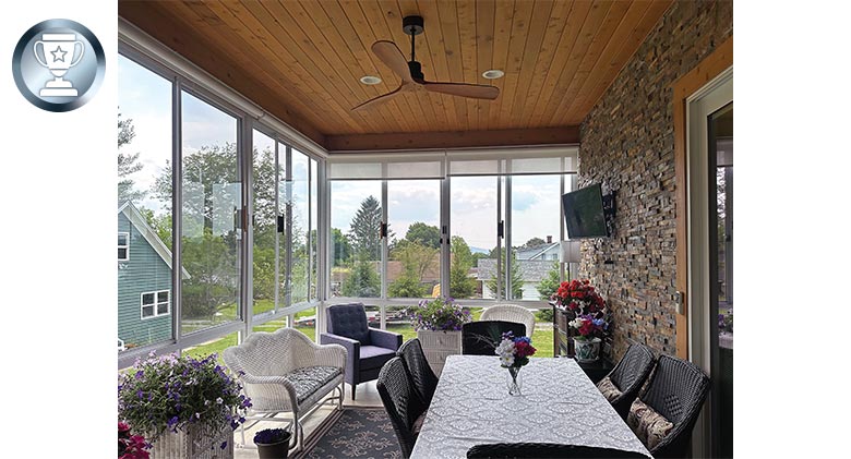 A dining table with tablecloth and some comfortable chairs and flowering plants decorate this glass porch enclosure.