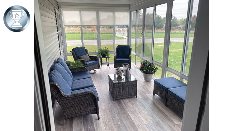 A glass-enclosed porch furnished with comfortable brown wicker chairs, blue cushions, ottomans, and side tables.