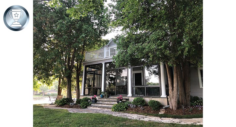 A historic Cape Cod house surrounded by trees with the lower front porch enclosed in glass and an open deck above.