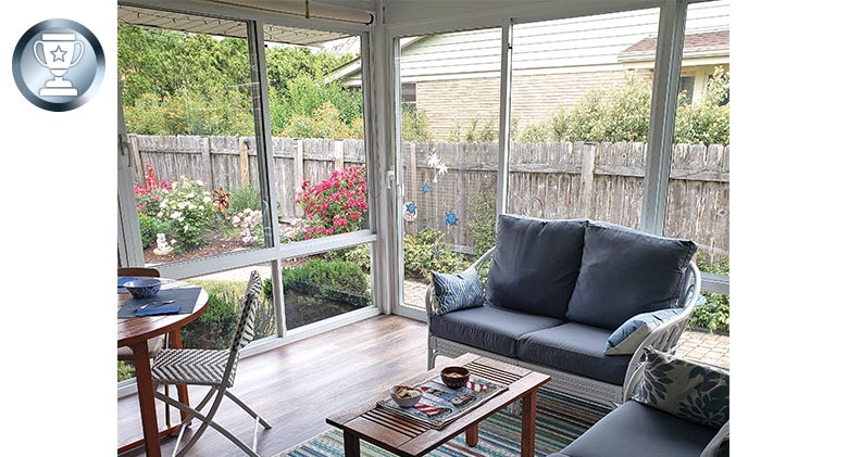 A glass-enclosed porch with seating and dining areas. Flowering gardens can be seen through the glass.