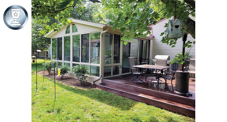 A beige sunroom with gable roof and two quarter-arch windows in the wing. An outside deck with table and chairs is adjacent.