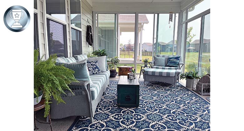 Sunroom interior with gray striped couch and chair, trunk as a coffee table, plants, and a blue patterned rug.
