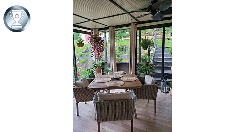 Inside of a glass-enclosed porch with a table and chairs, plants and ceiling fan. Outside, stairs leading up into the yard.