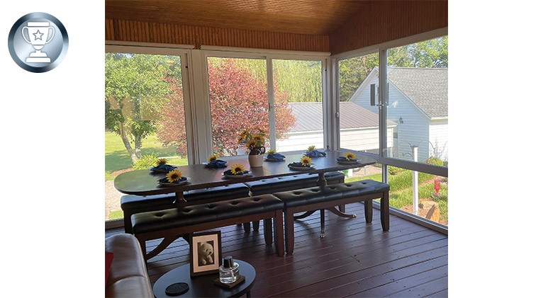 Interior of a second-floor enclosed porch showing a long table with place settings and benches, and a view of the yard.