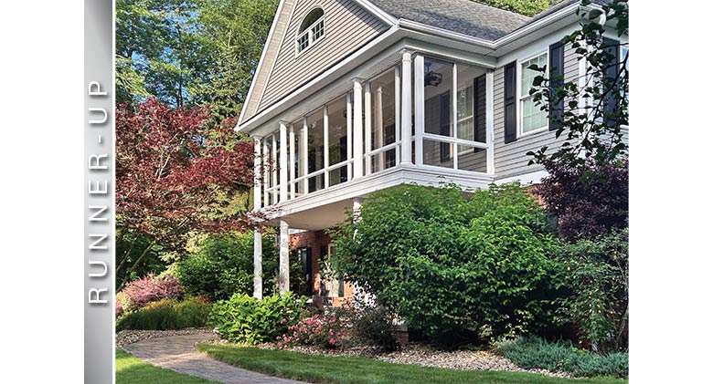 A second-story front porch enclosure on a tan home over an open front porch; nice landscaping surrounds the home.
