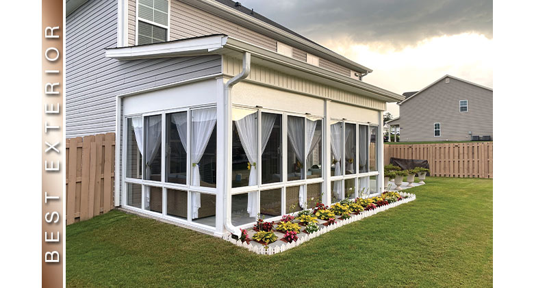 A covered patio enclosed with a white three-season room with tied-back curtains. A row of green and red plants in front.