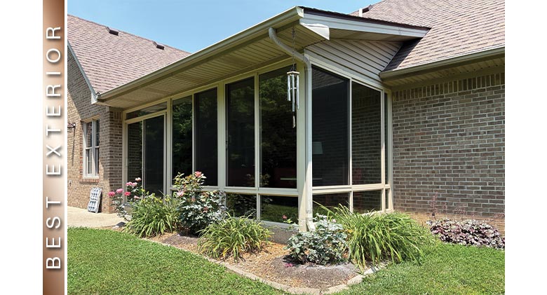 A covered patio enclosed with a tan three-season sunroom with glass knee wall and neat plantings in front.