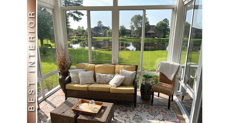 Yellow cushions and white pillows on brown wicker sofa in a glass sunroom with large vase, side tables, and side chair.