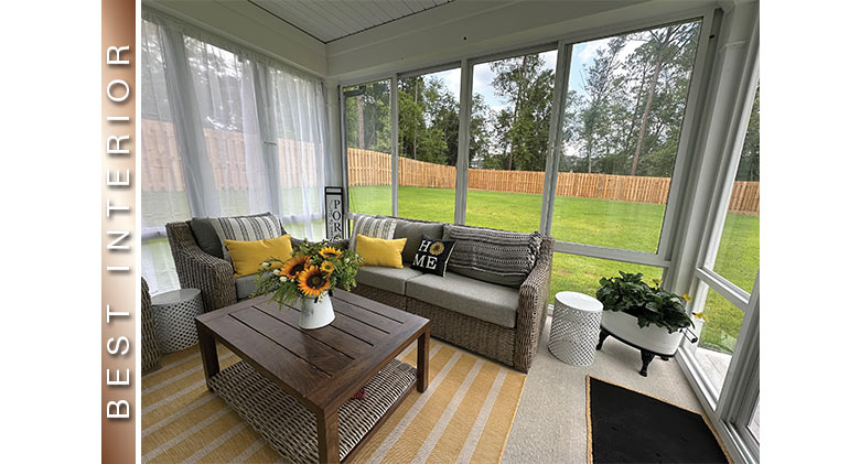 Sunflowers on wooden coffee table in glass sunroom with white sheers. Brown furniture with gray & yellow pillows & cushions.