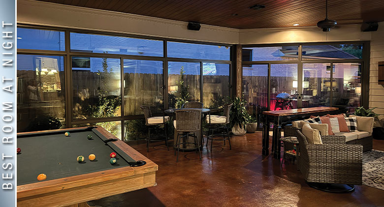 Nicely lit interior of a glass patio enclosure at night with a billiard table, dining table and comfortable seating area.