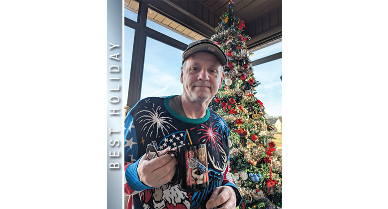 A man dressed in a patriotic holiday sweater with fireworks & Santa standing in front of a Christmas tree in a glass sunroom.