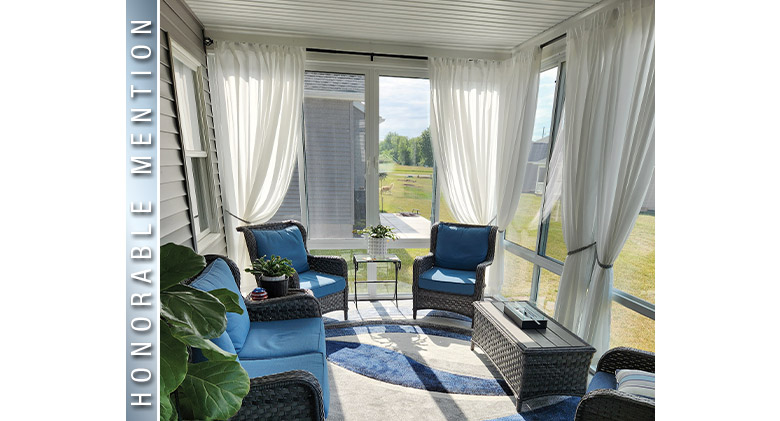 Two blue-cushioned wicker chairs and sofa in a glass patio enclosure with wicker coffee table and white tied-back curtains.