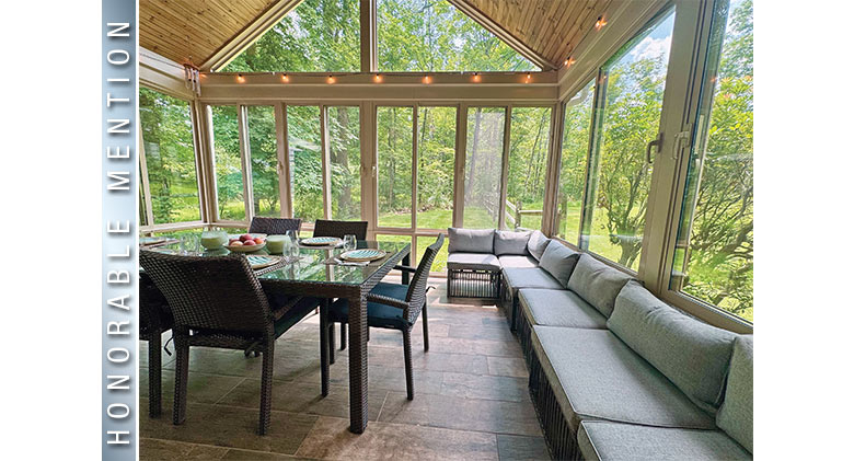 Interior of a glass sunroom with soaring ceiling and furnished with set dining table and chairs and a long bench sofa.