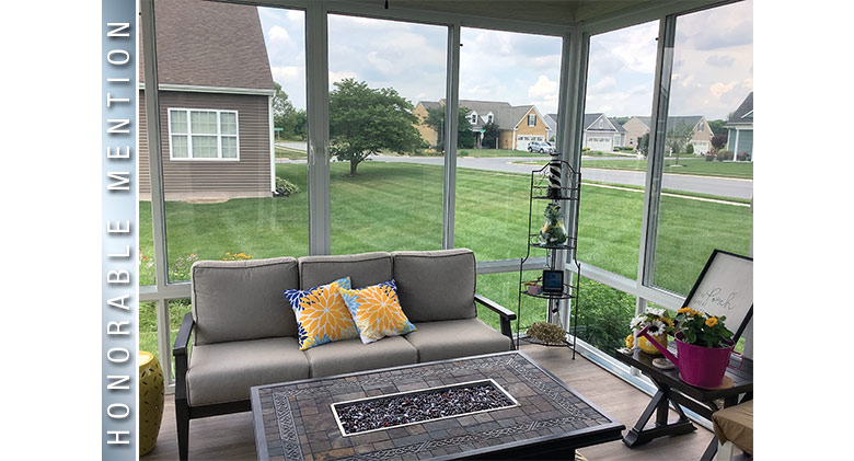 Inside of a glass patio enclosure with gray sofa & bright pillows, coffee table, & potted plants. View of the neighborhood.