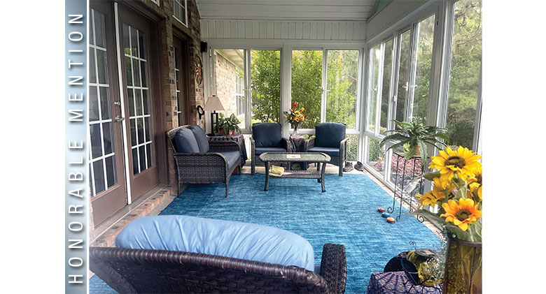Vases of sunflowers & plants decorate a glass porch enclosure with blue carpet and wicker furniture with blue cushions.