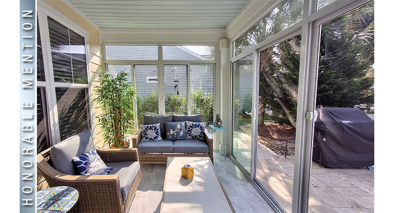 A glass patio enclosure with a chair facing the windows and a loveseat at the end of the room with a potted tree next to it.