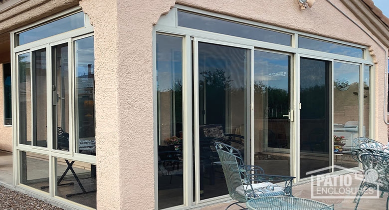 The exterior of a glass-enclosed corner patio on a stucco building with a table and chairs of iron on the patio outside.