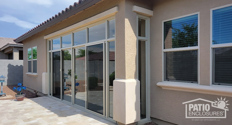 Patio enclosed with glass in stucco home with decorative pillars on both sides, paver patio in front and pot of blue flowers.