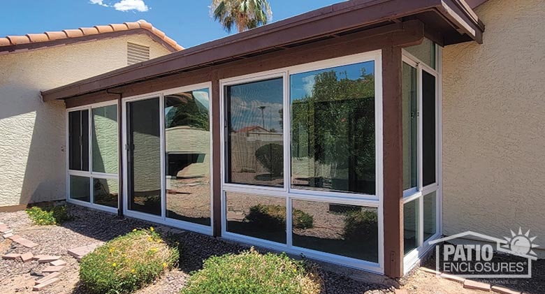 Glass patio enclosure with wood trim on a stucco home, sliding glass doors in the middle with gravel and plants in front.