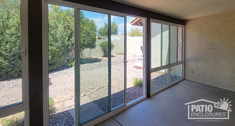 Interior of a glass-enclosed patio with white frame and wood trim overlooking backyard with gravel, bushes and small trees.