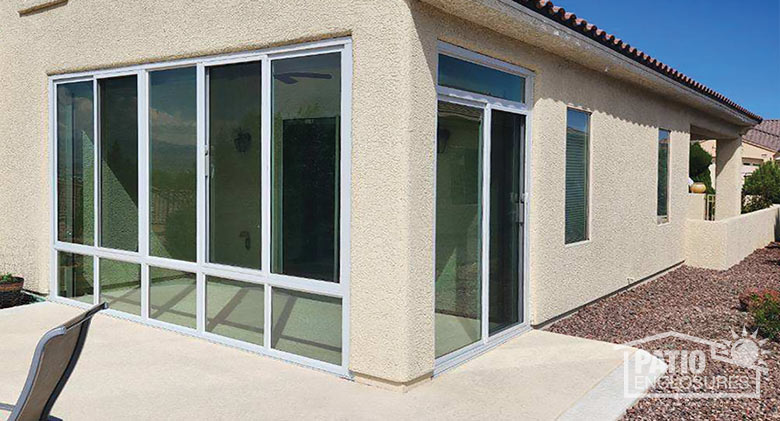 The exterior of a glass-enclosed corner patio on a beige stucco home with tile roof. A low stucco wall in the background.