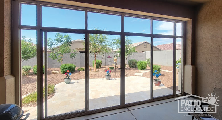 Interior of a glass-enclosed patio overlooking backyard with paver patio, potted plants, stepping stones, bushes and trees.