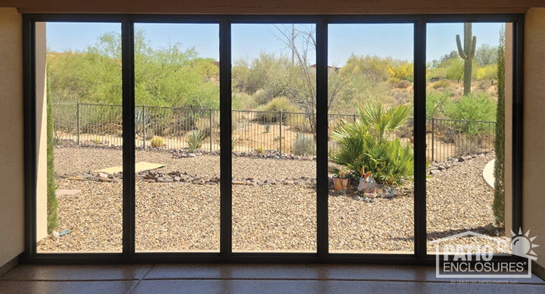 Interior view from a glass patio enclosure with a view of backyard gravel and plants, a fence and scrubby hillside.