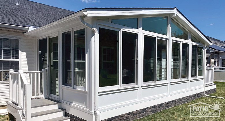: A white sunroom with gable roof, solid knee wall and glass wings. Three stairs on the left lead to sliding patio door.